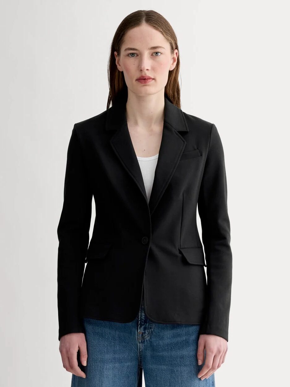 A person with straight brown hair wears a black blazer over a white top and blue jeans, standing against a plain light background.