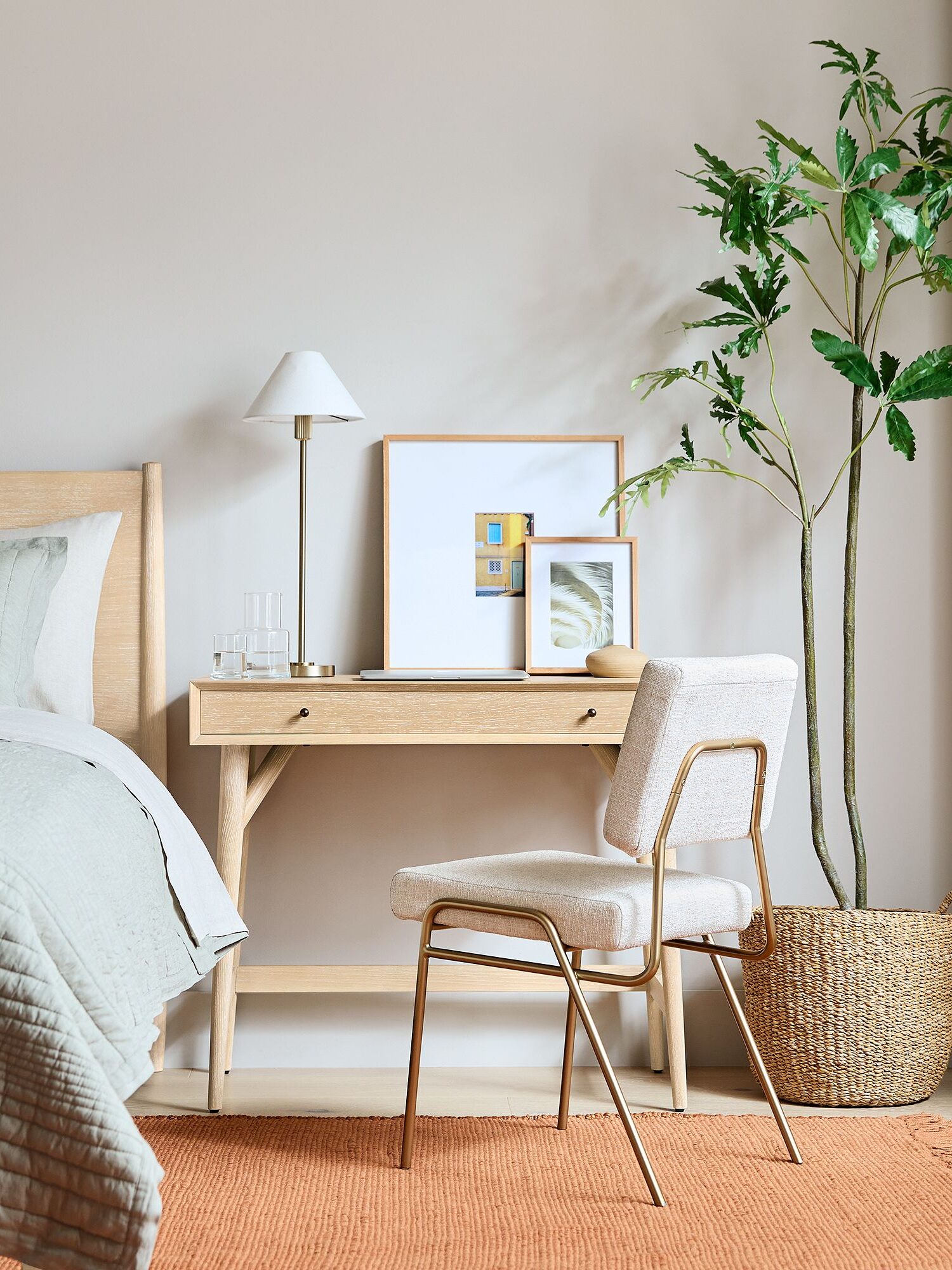 A minimalist bedroom with a wooden bed, desk, upholstered chair, potted plant, framed art, lamp, and orange rug on a light floor.