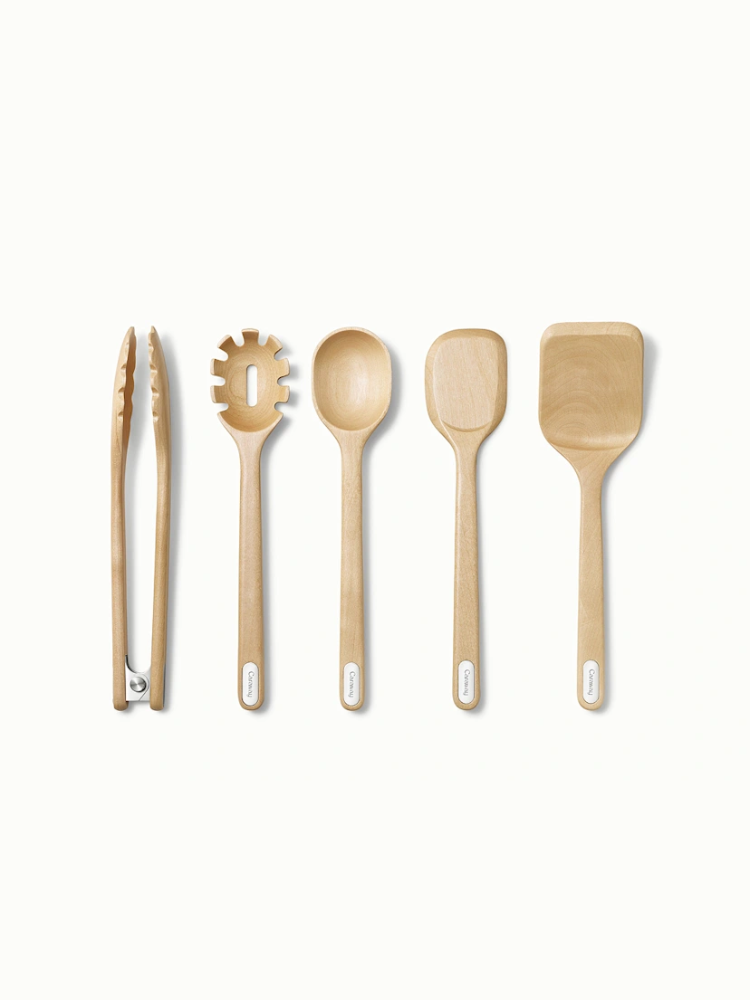 A set of five wooden kitchen utensils, including tongs, a pasta server, a spoon, a spatula, and a flat turner, arranged side by side on a white background.