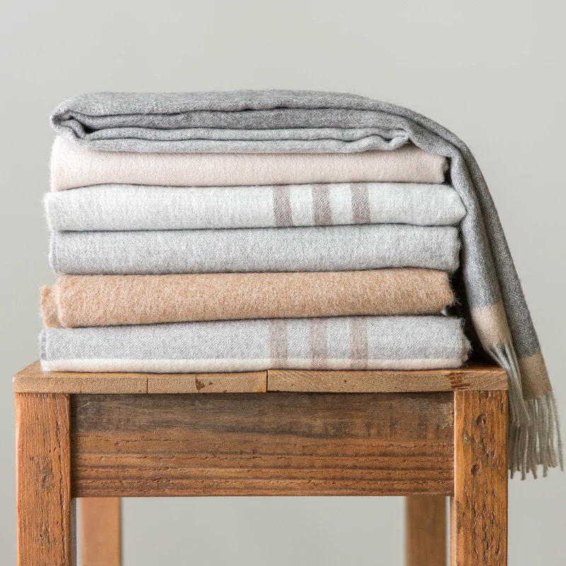 A stack of neatly folded wool blankets in neutral colors sits on a wooden stool against a plain background.