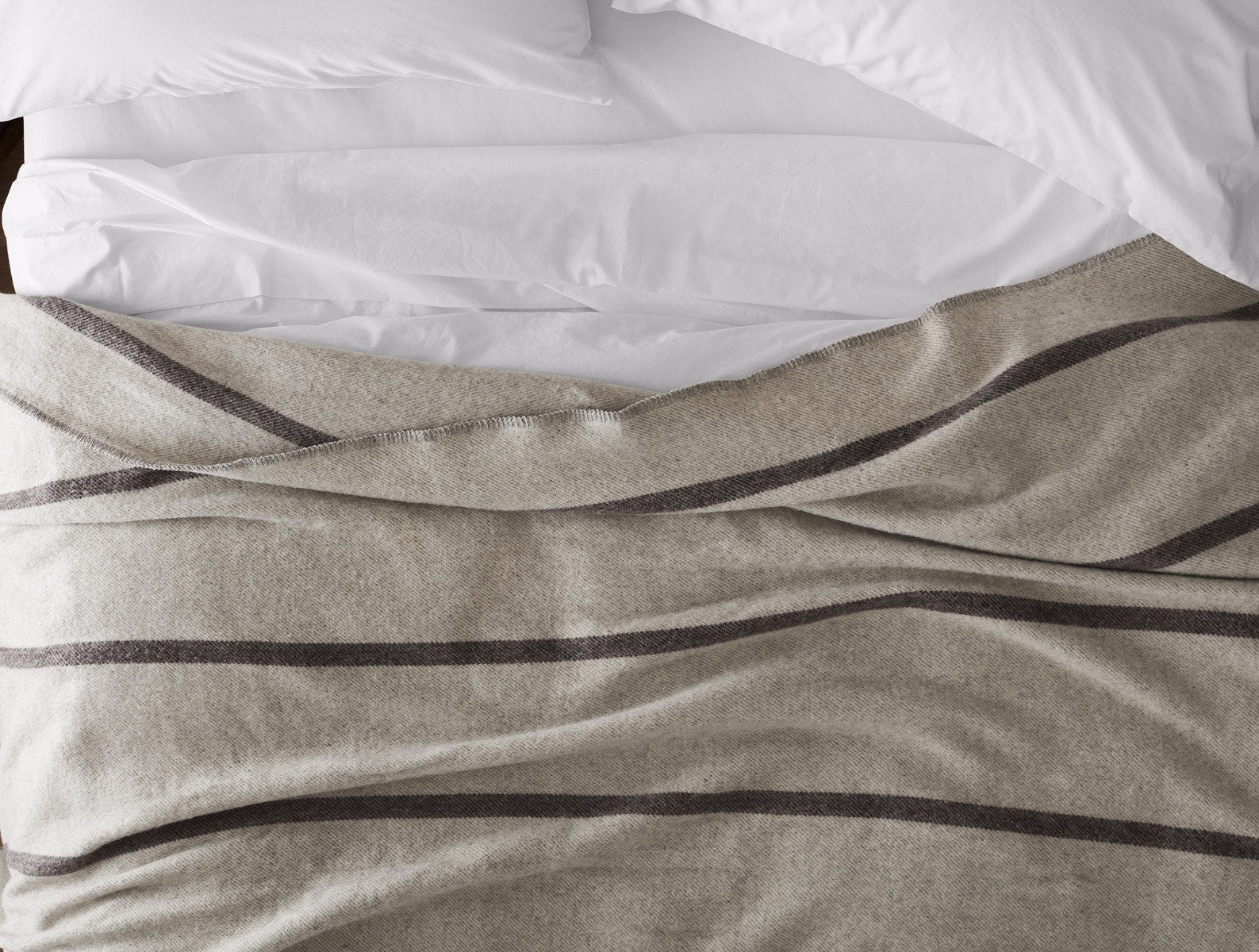 A neatly made bed with white sheets and pillows, partially covered by a beige blanket with dark horizontal stripes.