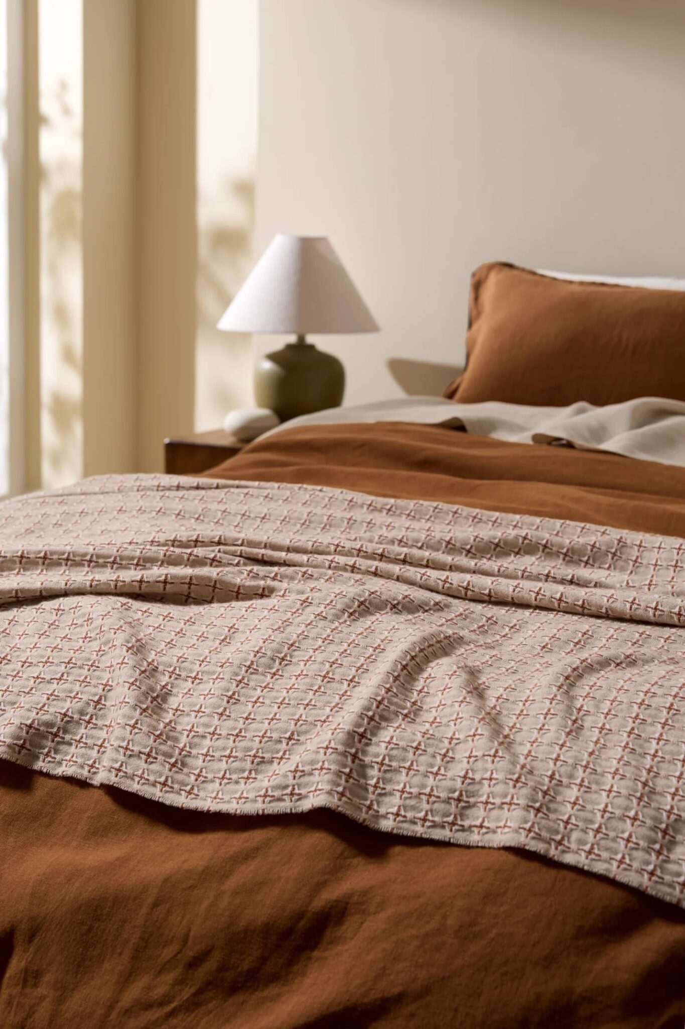 A neatly made bed with brown and beige bedding, a textured blanket, and a bedside table holding a lamp, set in a sunlit room.