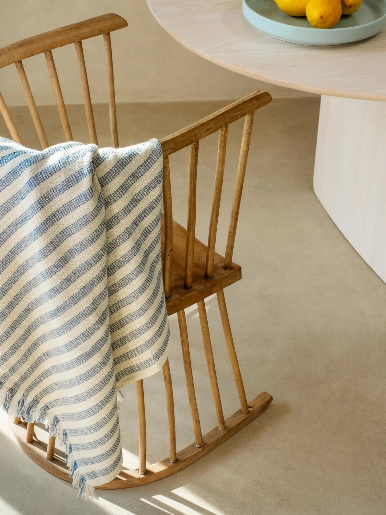 A wooden rocking chair with a blue and white striped blanket draped over it sits next to a round table with a bowl of lemons.