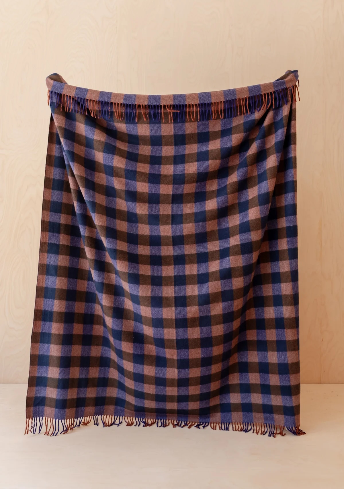 A wool blanket with a blue and brown checkered pattern is draped over a wooden surface against a light beige background.