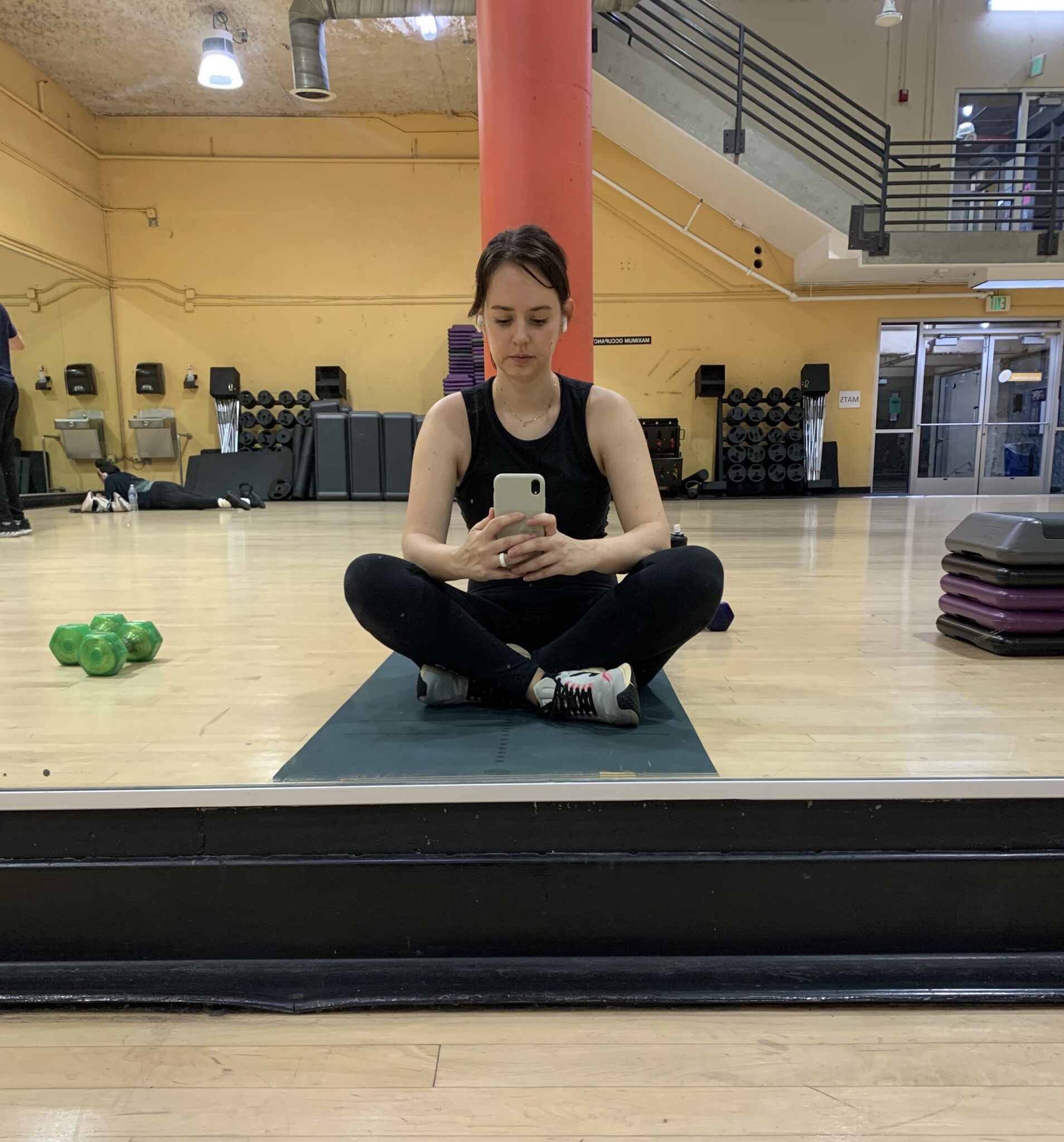 A person in workout attire sits cross-legged on a mat in a gym, taking a selfie in the mirror, with weights and equipment visible in the background.
