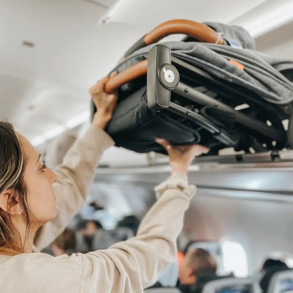 A person places a folded stroller into an overhead compartment on an airplane.