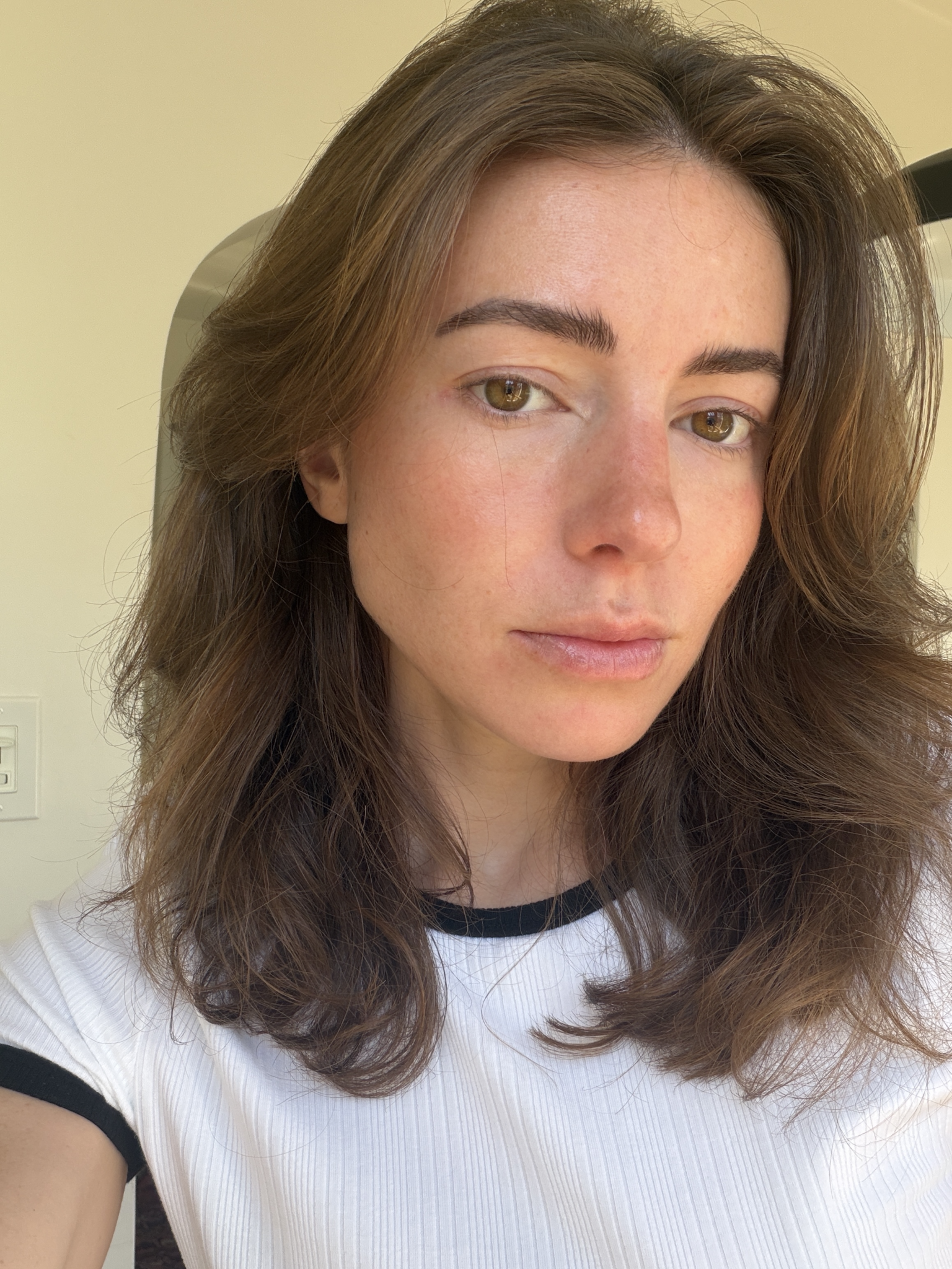 A person with long brown hair and light skin, wearing a white shirt with black trim, looks at the camera in natural lighting.