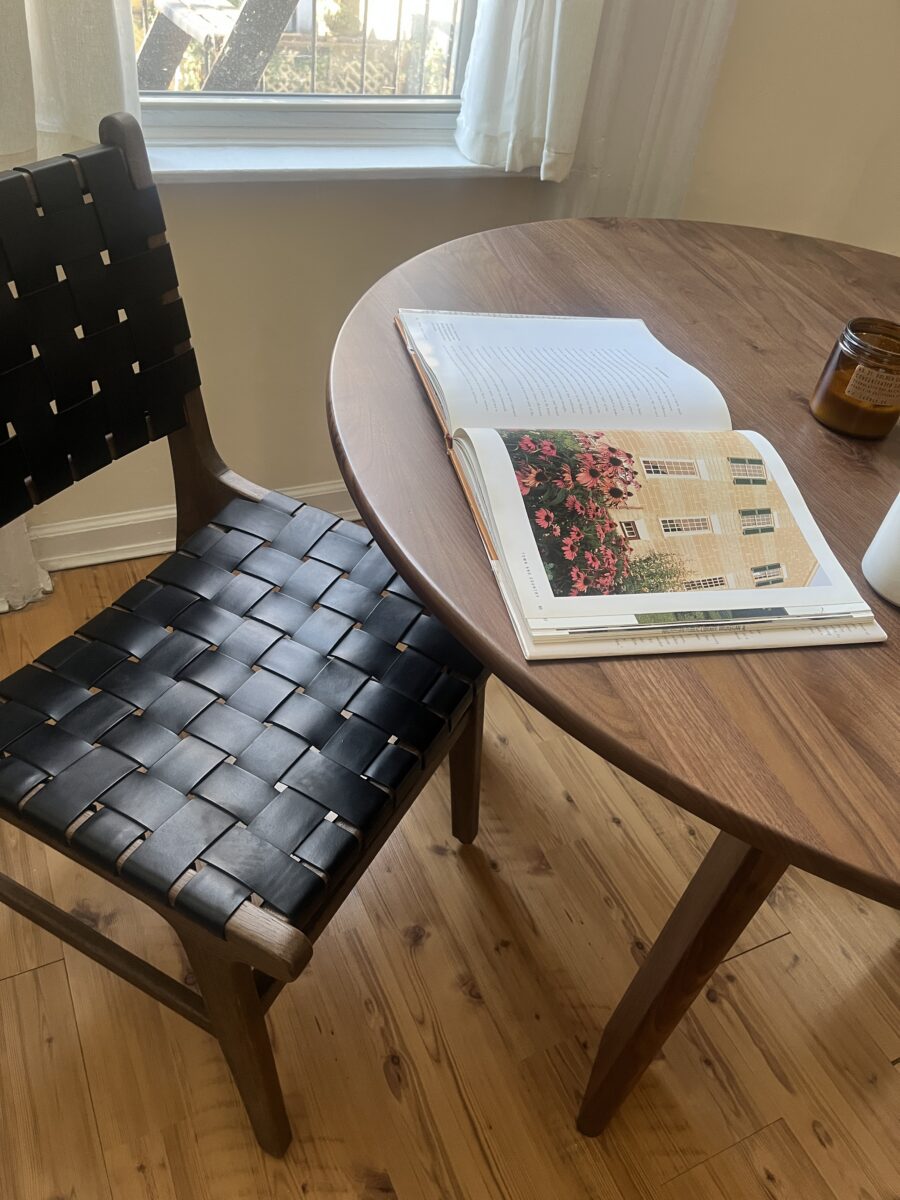 A wooden round table with an open book, a brown glass jar candle, and a woven black chair next to it on a wooden floor by a window with sheer curtains.