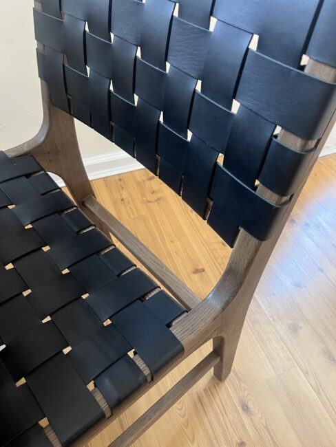 A wooden chair with a seat and backrest made of interwoven black straps, placed on a light-colored wooden floor.