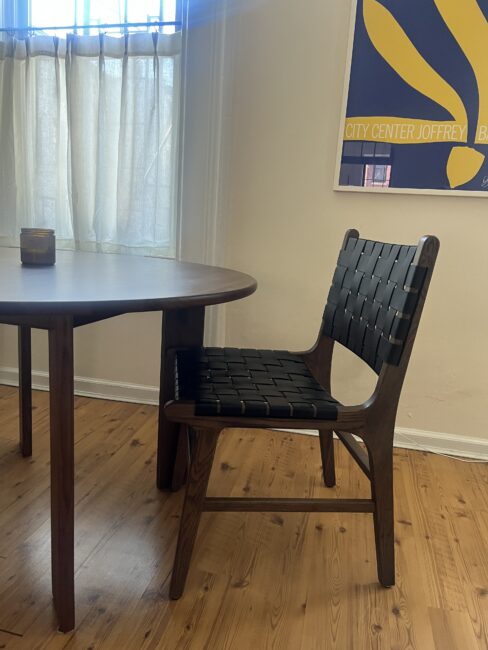 A wooden dining table with a candle on top sits next to a black woven chair on a wood floor; a window and framed poster are in the background.