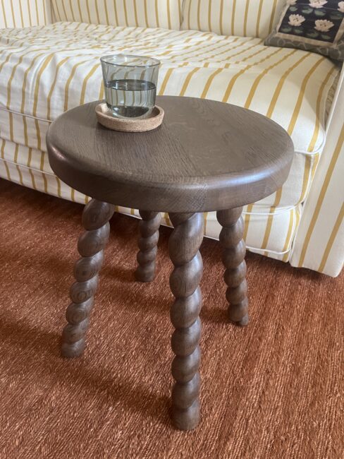 A dark wooden stool with spiral legs holds a glass of water on a coaster, positioned in front of a yellow and white striped sofa on a brown carpet.