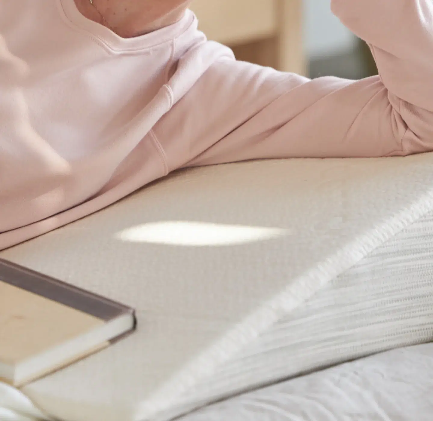 A person wearing a light pink long-sleeve shirt rests their arm on a layered mattress, with a closed book nearby on the bed.