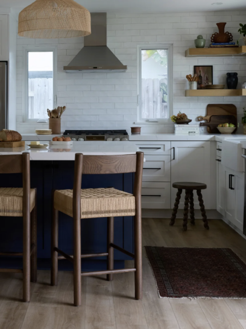Modern kitchen with white cabinets, a navy island, wooden barstools, open shelves, stainless steel range hood, and a rug on light wood flooring.