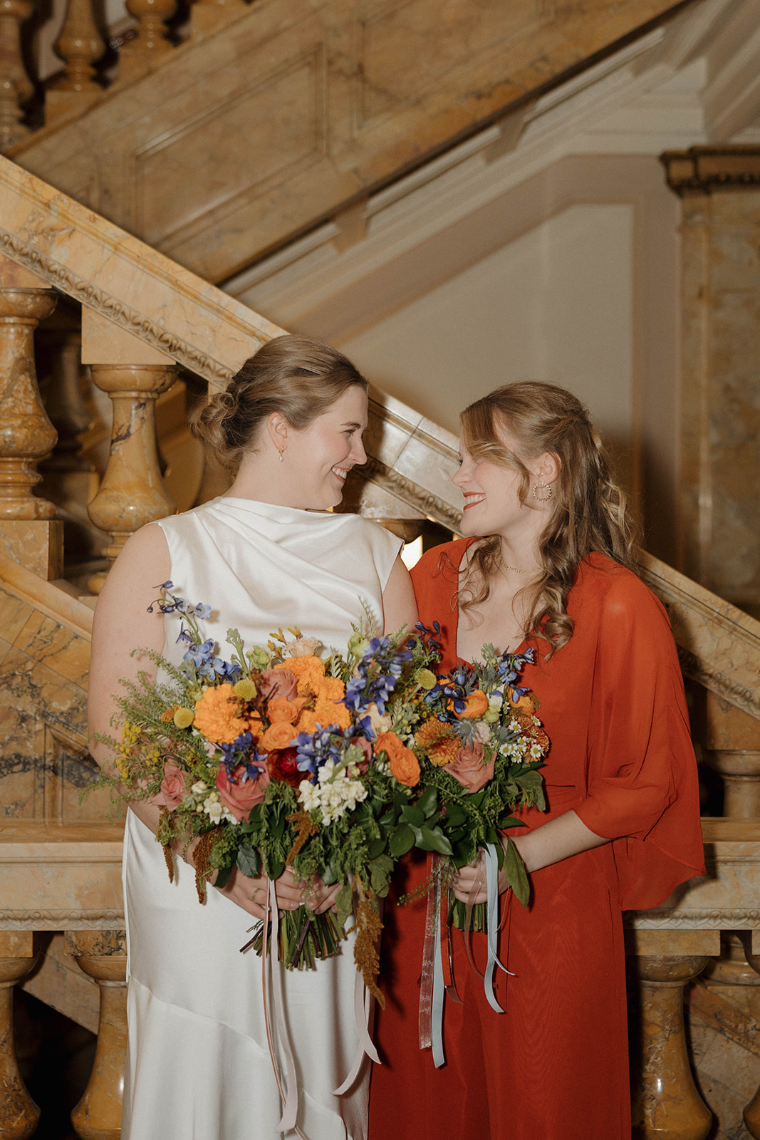 Two women stand on marble stairs, each holding a large bouquet of colorful flowers, looking at each other and smiling.