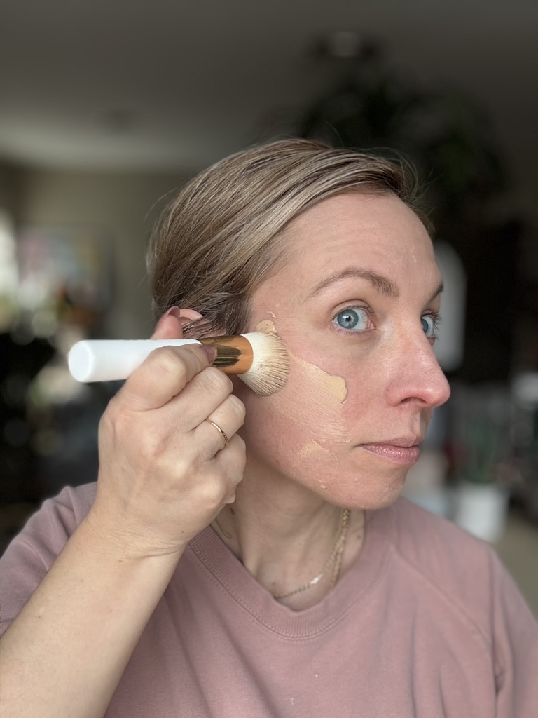 A person applies Ilia liquid foundation to their cheek with a makeup brush, holding the brush in their right hand and looking slightly upward.