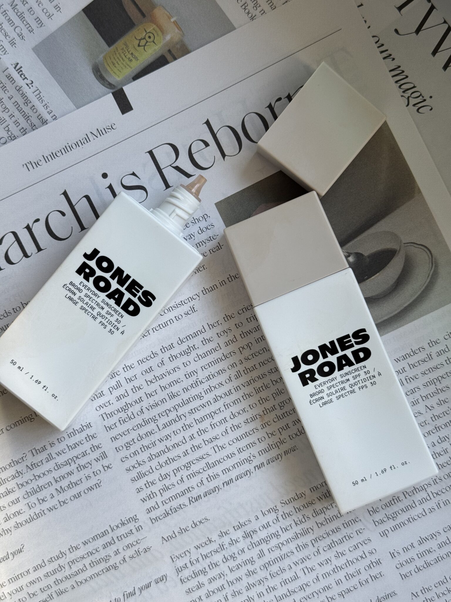 Two Jones Road foundation bottles, one standing with the cap off and one lying down, are placed on a newspaper—reminiscent of the natural beauty ethos seen in Ilia products. One bottle reveals product at the nozzle.