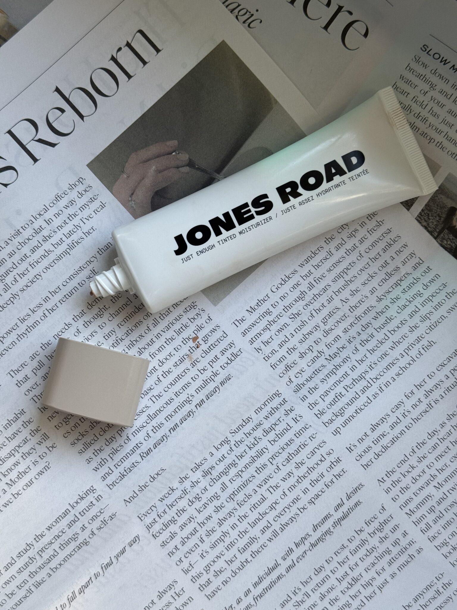 A tube of Jones Road Tinted Moisturizer with its cap off rests on an open newspaper, beside an Ilia product.
