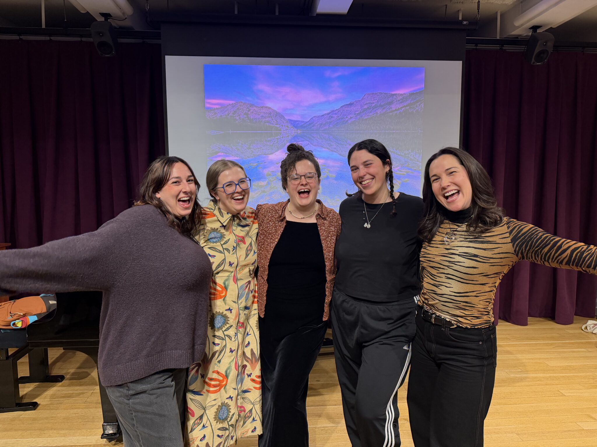 Five people stand arm-in-arm in front of a projected scenic mountain image, smiling and posing for the camera in a room with purple curtains.