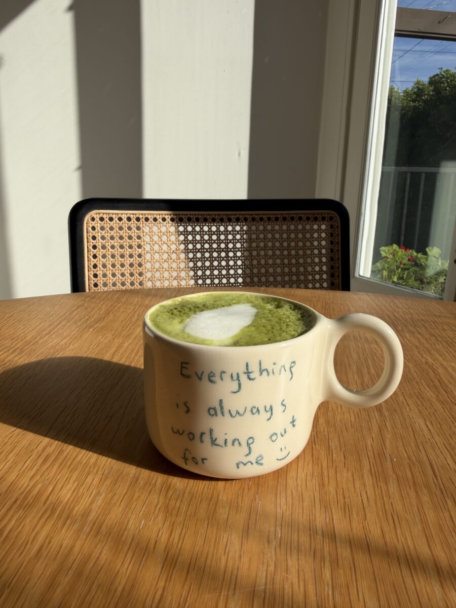 A cup of green matcha latte with heart-shaped foam sits on a wooden table. The mug reads, "Everything is always working out for me :)" A window and chair are in the background.