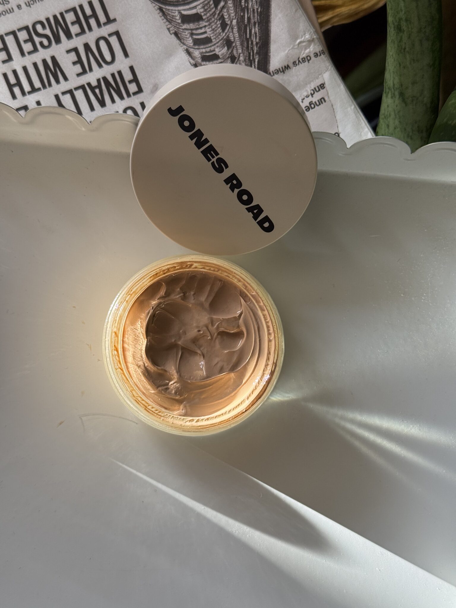 An open container of Jones Road makeup sits on a white surface with its lid beside it, exposing the beige product inside—reminiscent of minimalist Ilia beauty essentials.