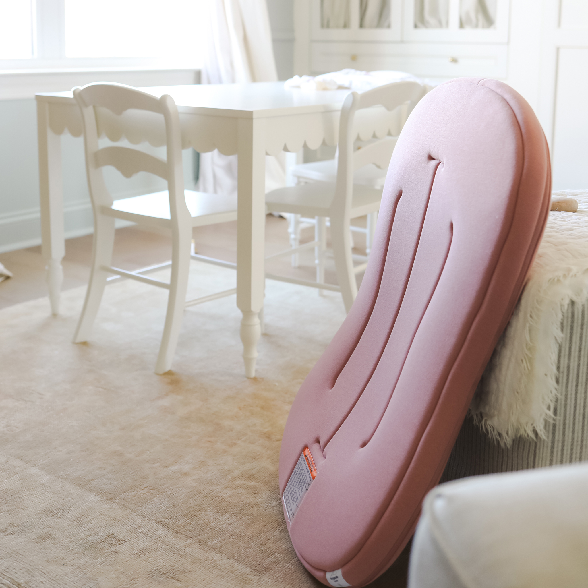 A pink baby lounger is propped against a beige sofa in a bright room with a white table and chairs in the background.