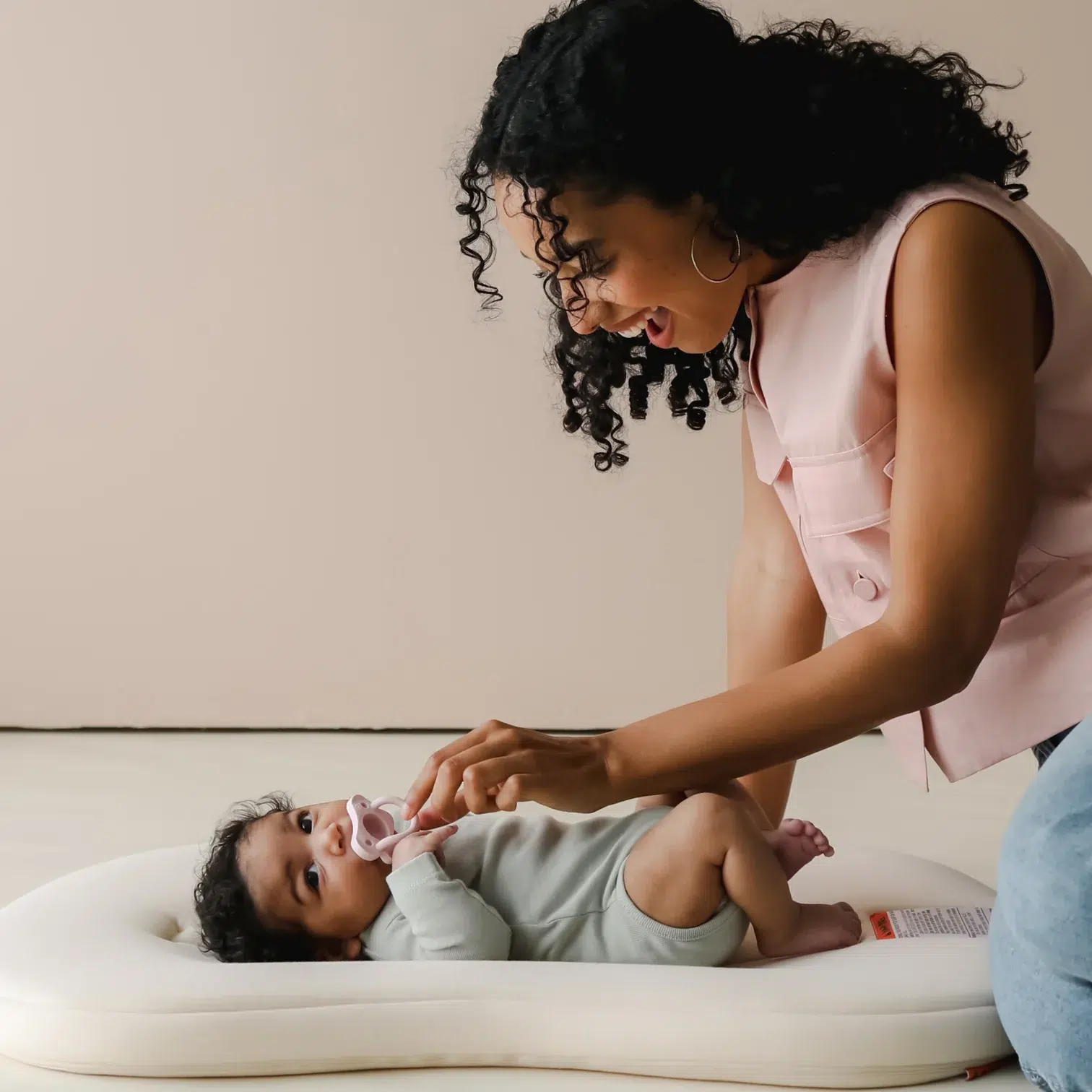 An adult woman smiles while holding a pacifier for a baby lying on a cushioned mat.