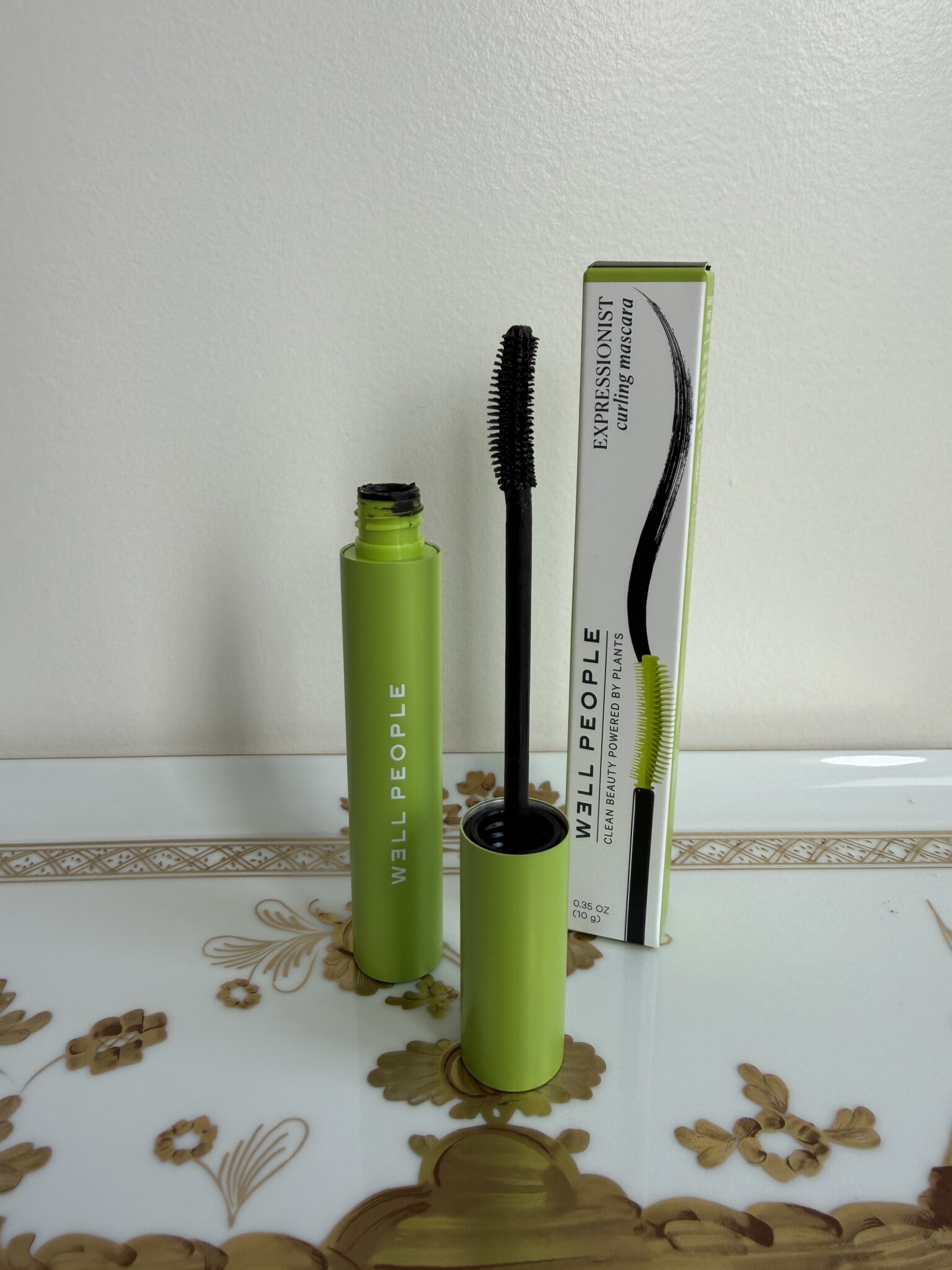 A green tube of Well People mascara with the wand exposed, standing next to its product box on a decorative tray.