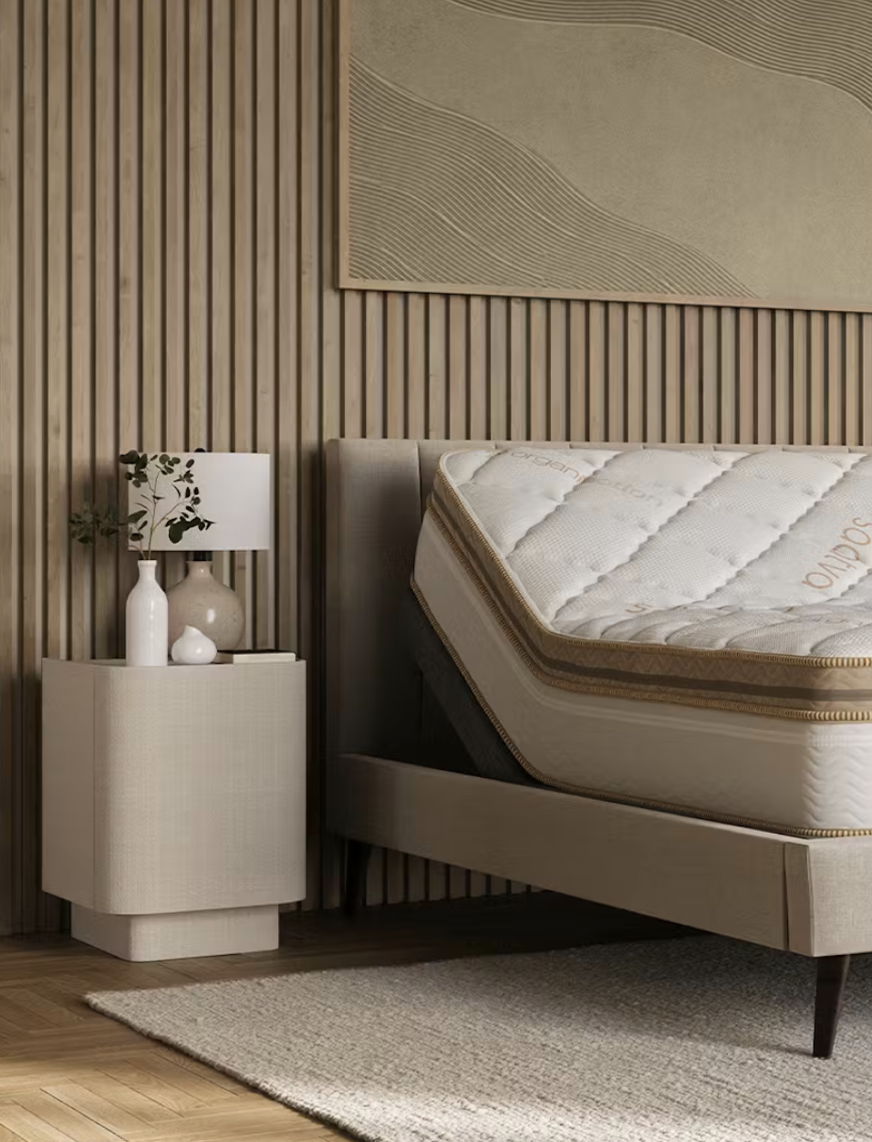 A bedroom with a modern beige bed frame, mattress propped up, nightstand with a lamp and vases, wood panel wall, abstract art, and a textured rug.