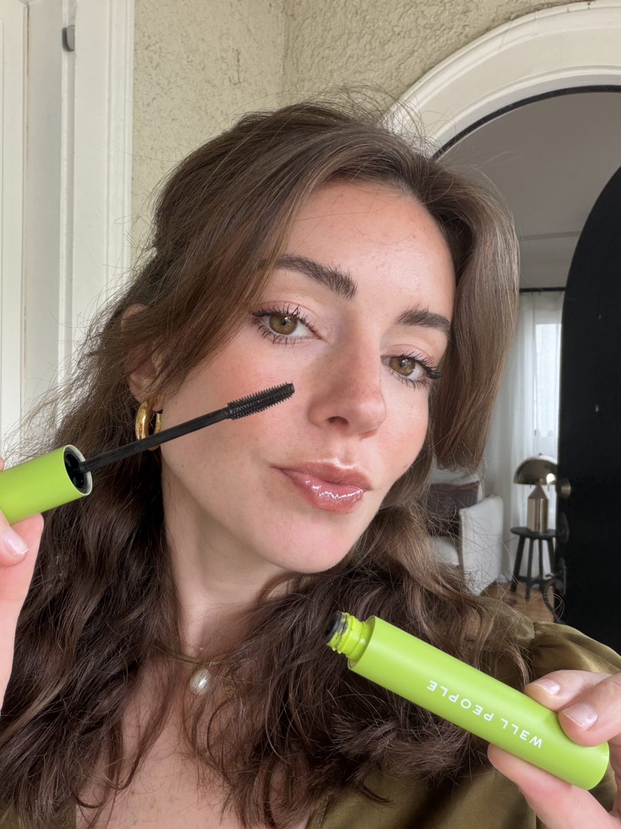 A woman with wavy brown hair applies mascara to her eyelashes using a wand. She is holding a green tube labeled “WELL PEOPLE.”.