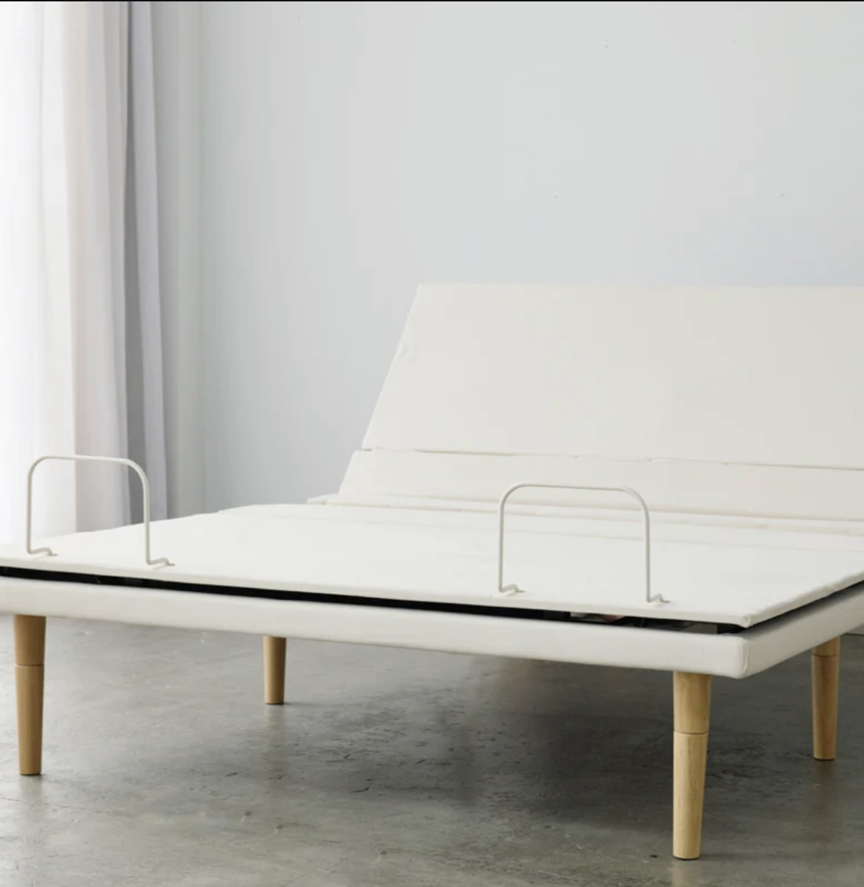 A white adjustable bed frame with light wooden legs is shown in a minimalist room with concrete floors and light curtains.
