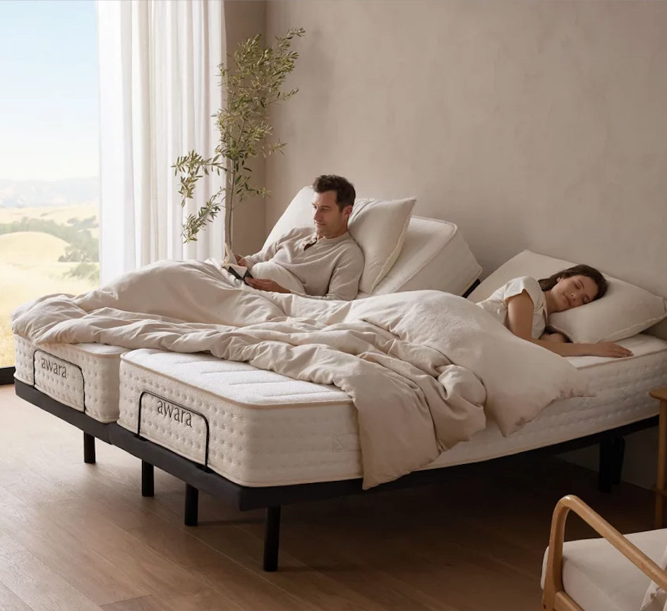 A man sits up reading while a woman sleeps on an adjustable split mattress bed in a bright, modern bedroom.
