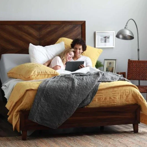 Two people are sitting up in bed together under yellow and gray bedding, looking at a tablet. There is a lamp, framed pictures, and a woven chair in the background.