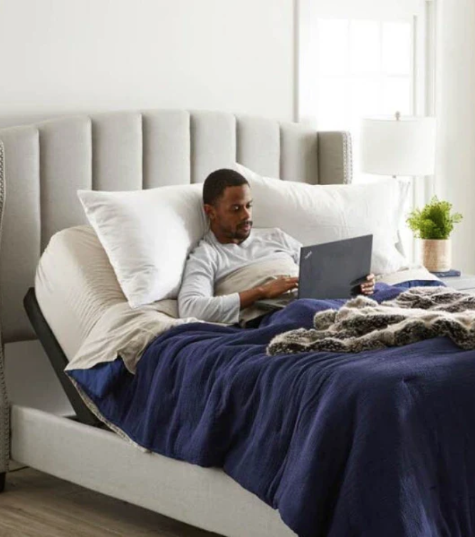A person reclines on a bed with an adjustable backrest, using a laptop. The bed has blue bedding, a white pillow, and a throw blanket; a lamp and plant sit on a nearby nightstand.