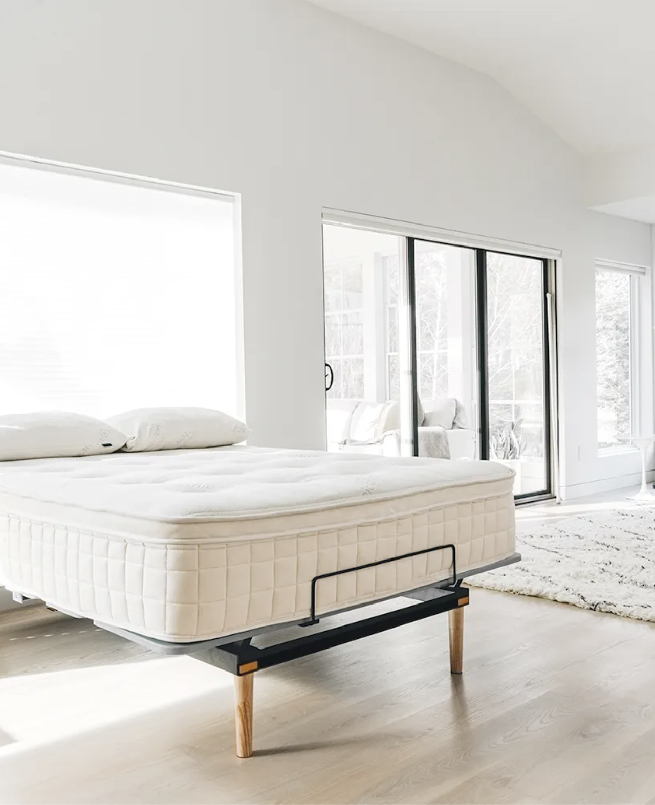 A neatly made bed with a white mattress on a modern bed frame stands in a bright room with large windows and light wooden floors.