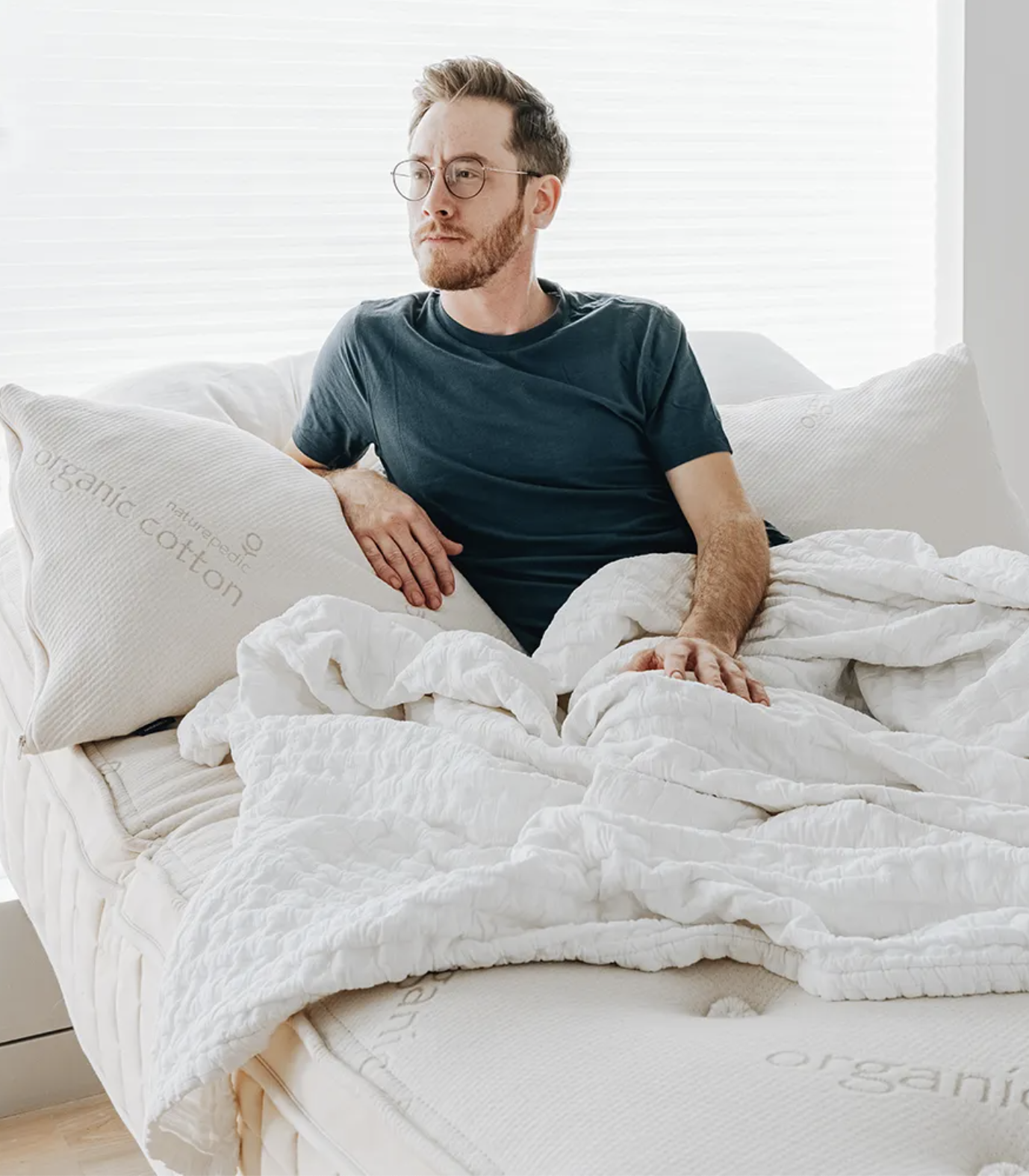 A man with glasses and a beard sits upright in bed, leaning on pillows with "organic cotton" labels, surrounded by white bedding in a bright room.