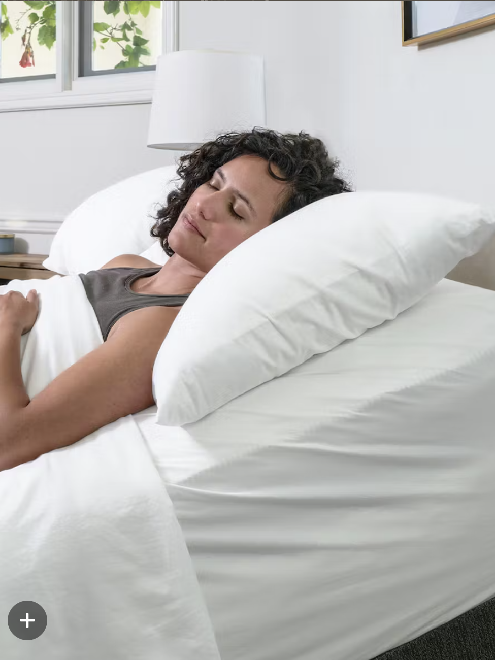A person with curly hair lies on a white adjustable bed, propped up by pillows, appearing relaxed and at rest in a bright bedroom.