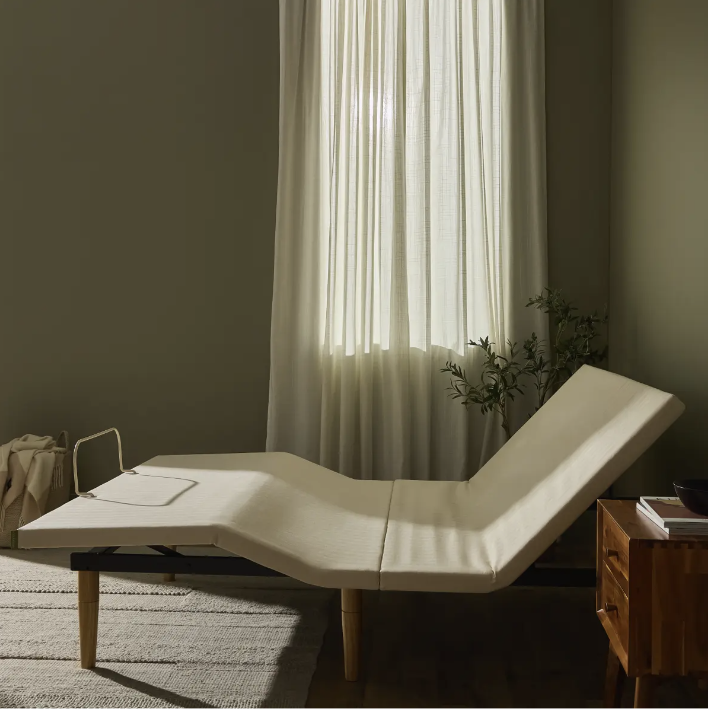 An adjustable bed in a partially upright position sits in a softly lit bedroom with light curtains, a wooden nightstand, and a plant nearby.