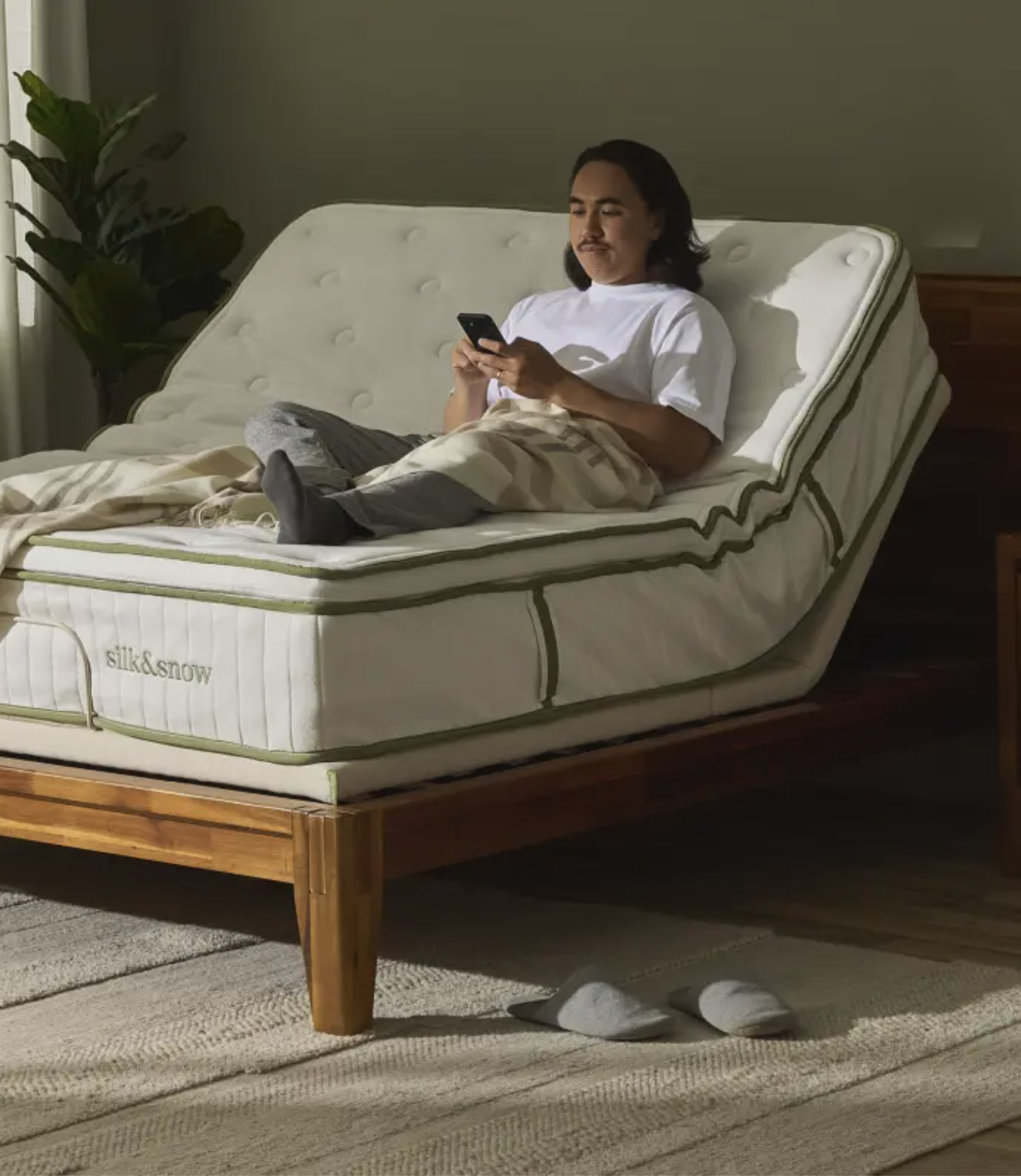 A person sits on an adjustable bed, using a smartphone. The bed is partially elevated, and slippers are on the floor nearby.