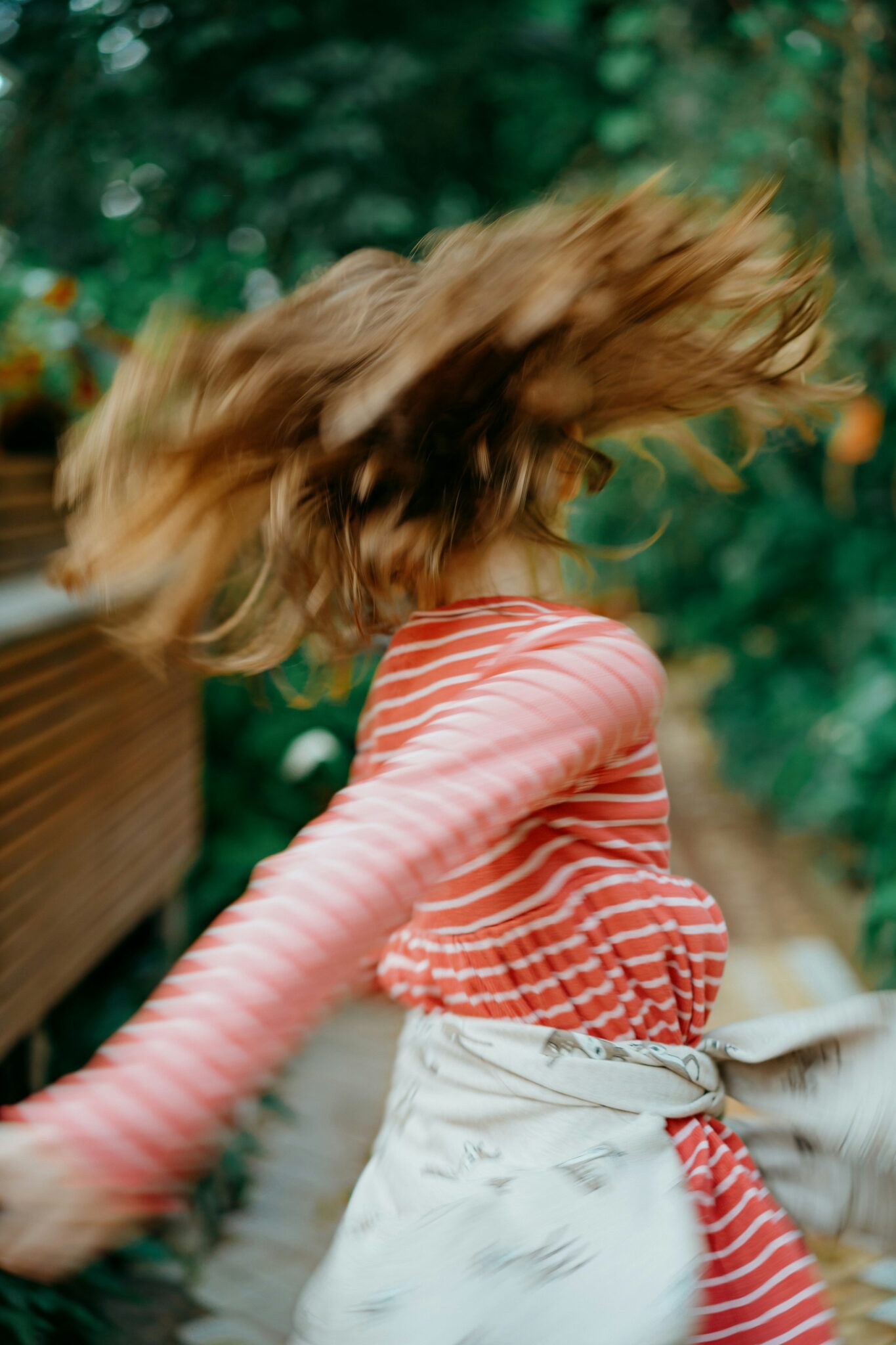 Person in a red and white striped top spinning outdoors, with hair and arms in motion, creating a blurred effect. Lush greenery is visible in the background.