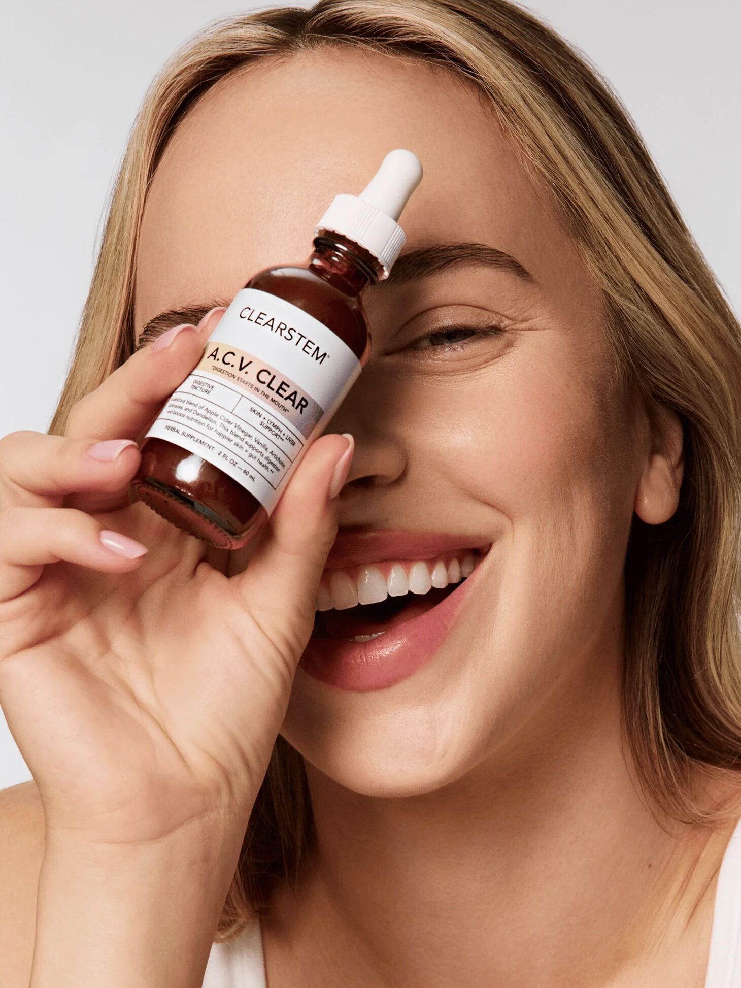 A smiling woman holds a dropper bottle labeled "CLEARSTEM A.C.V. CLEAR" up to her eye against a plain background.