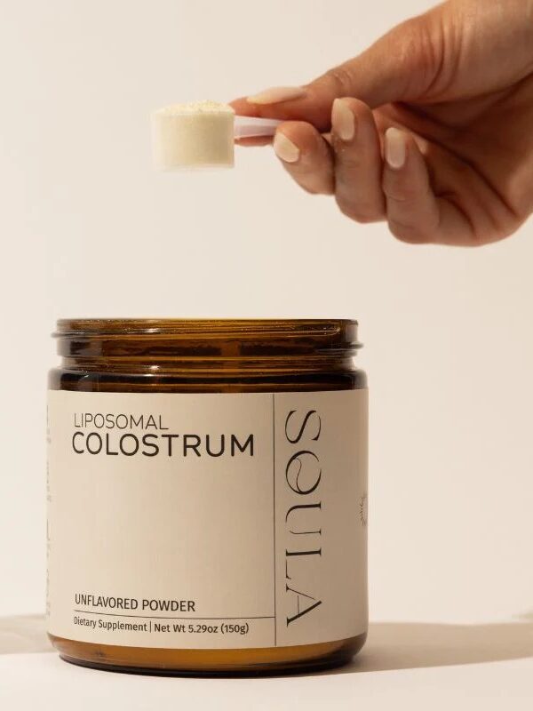 A hand holds a scoop of powder above an open jar labeled "Liposomal Colostrum Unflavored Powder Dietary Supplement" from the brand SOULA.