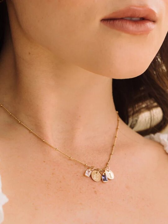 A close-up of a woman wearing a gold necklace with three small, engraved pendants, shown against a white blouse.
