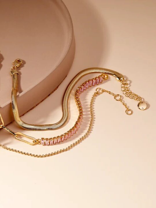 Four gold bracelets with varying chain styles, including beaded, paperclip, snake, and a pink gem-accented design, arranged on a pale pink surface.