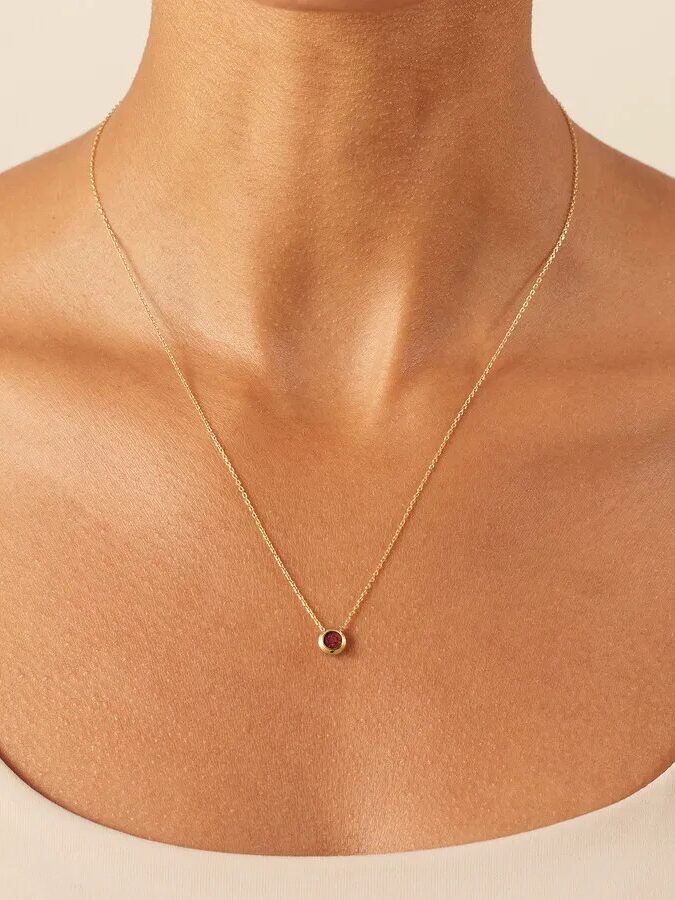 A close-up of a person wearing a delicate gold necklace with a small circular pendant featuring a red gemstone, against a neutral background.