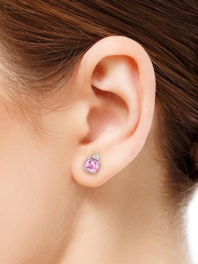 Close-up of a woman's ear wearing a small stud earring with a pink gemstone and a small clear stone above it.