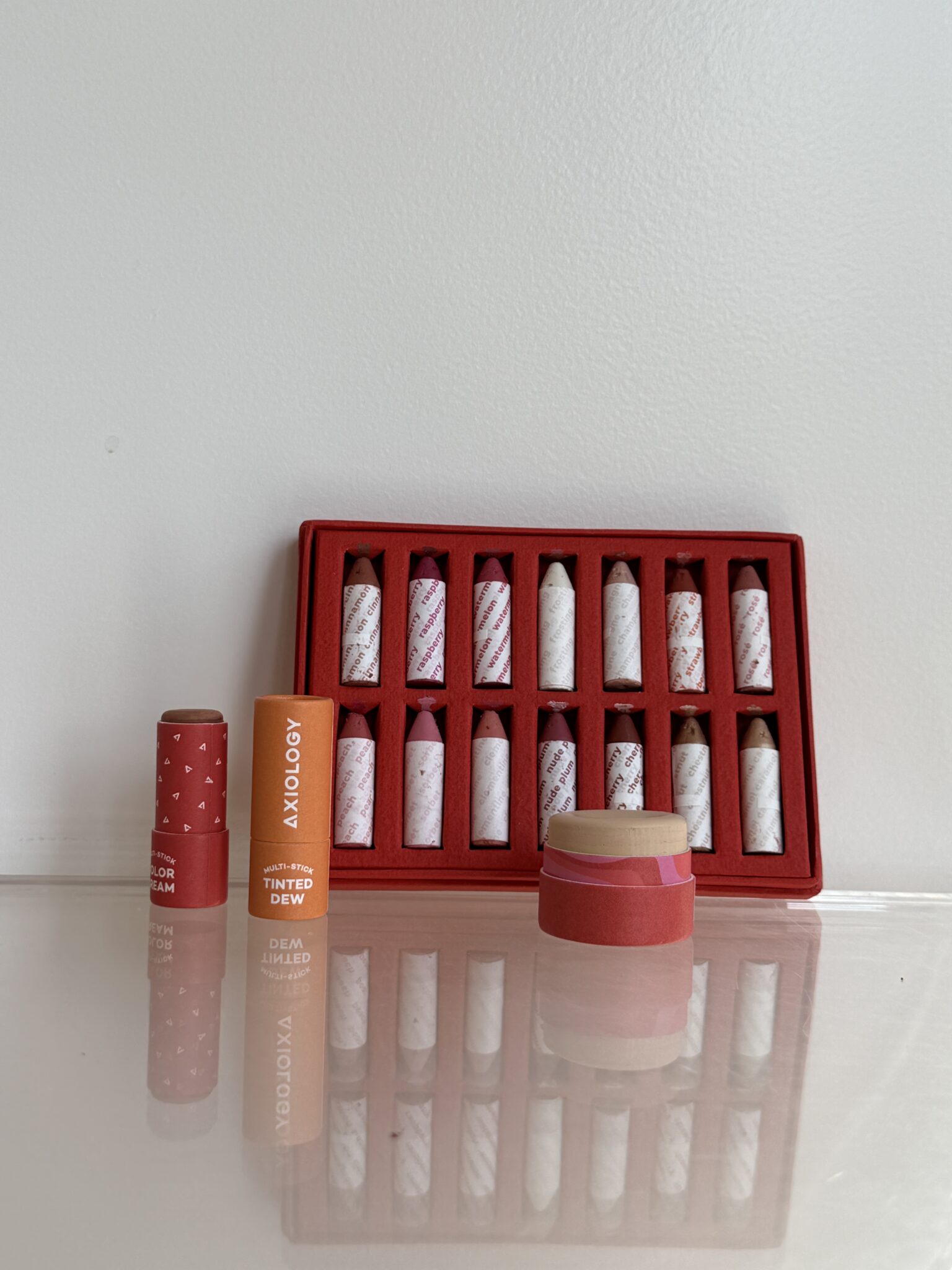 A set of ten lip balms in a red tray, with three individual lip balm tubes and one open container displayed in front on a reflective surface against a white background.