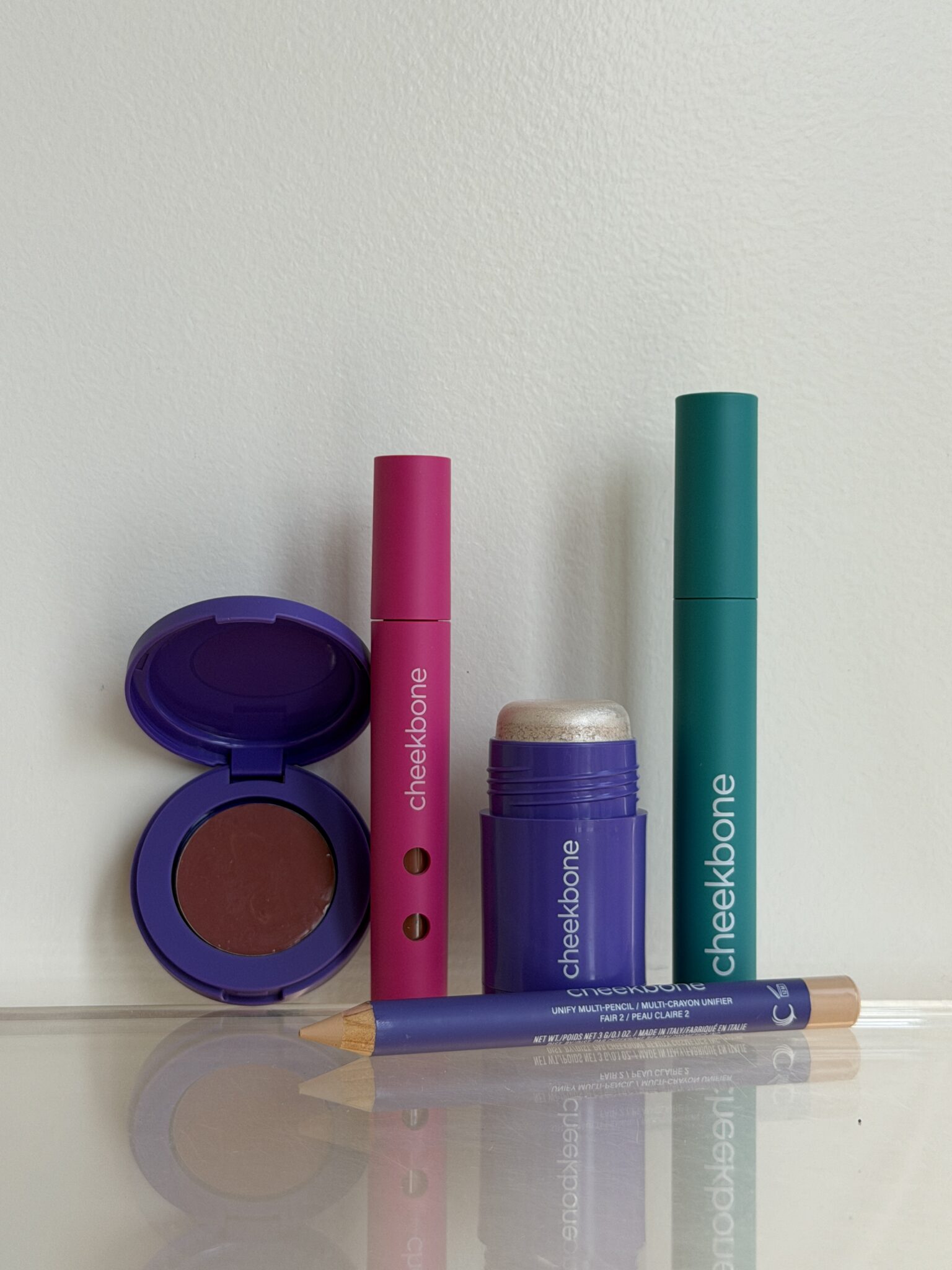 Four makeup products, including a compact, a stick, a pencil, and a cylindrical tube, are arranged upright against a plain white background.