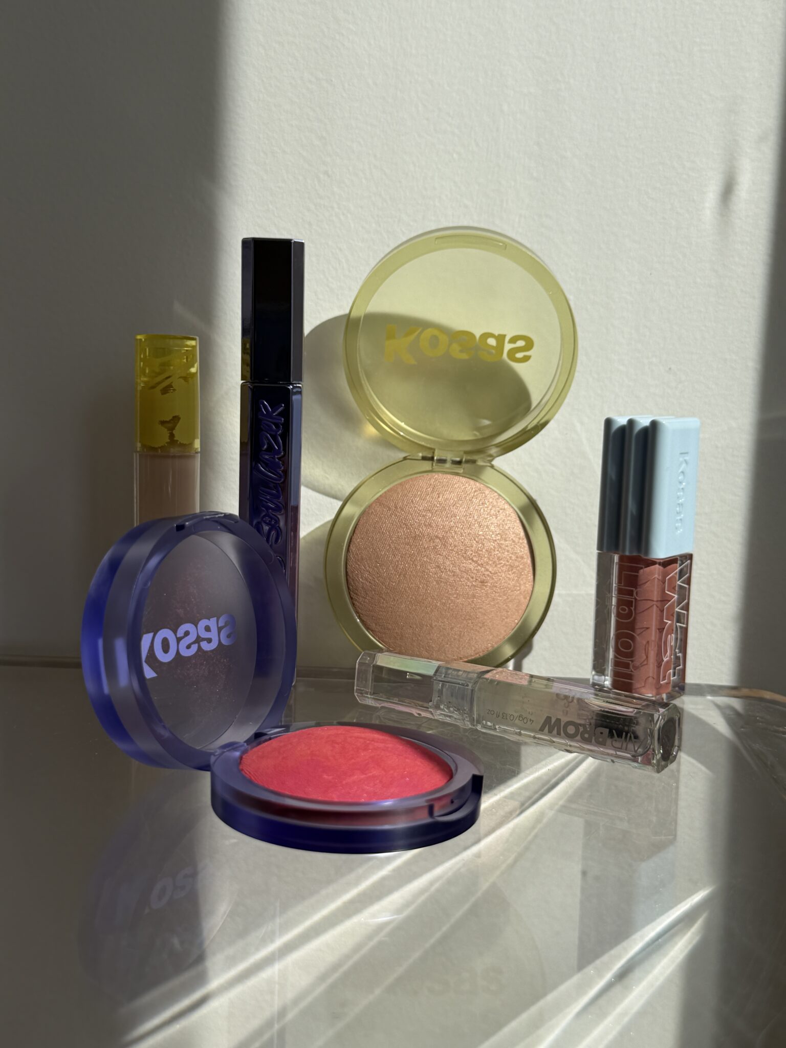 Assorted Kosas makeup products arranged on a reflective surface in sunlight, including blush, highlighter, lip gloss, and mascara.