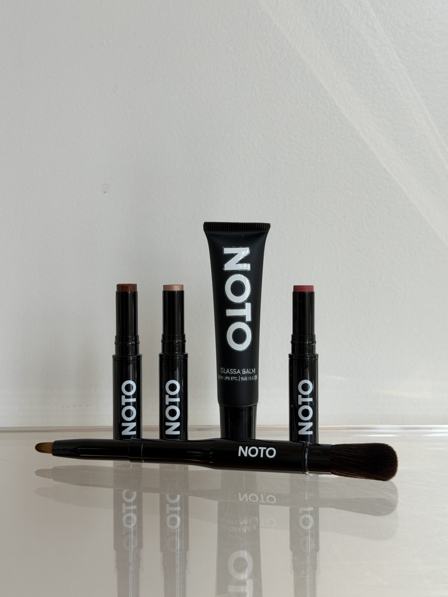A NOTO makeup set including three stick products, one tube, and a dual-ended brush arranged neatly on a reflective surface.