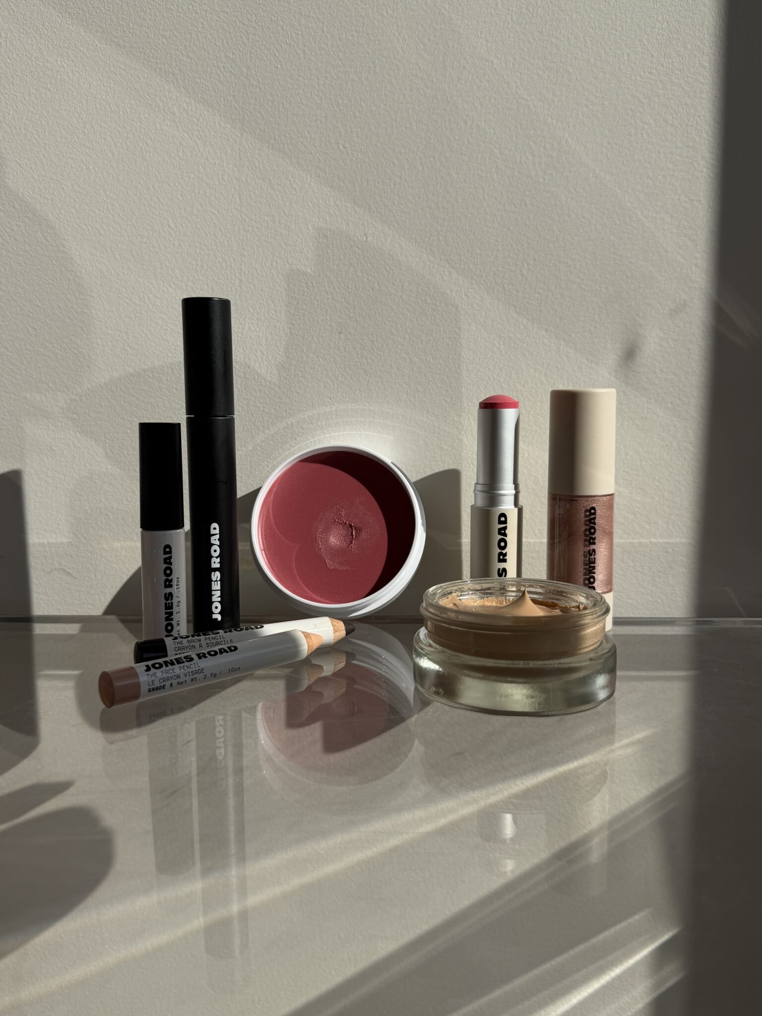 A selection of Jones Road makeup products, including lipsticks, a blush, a compact, and two makeup sticks, arranged on a sunlit surface.